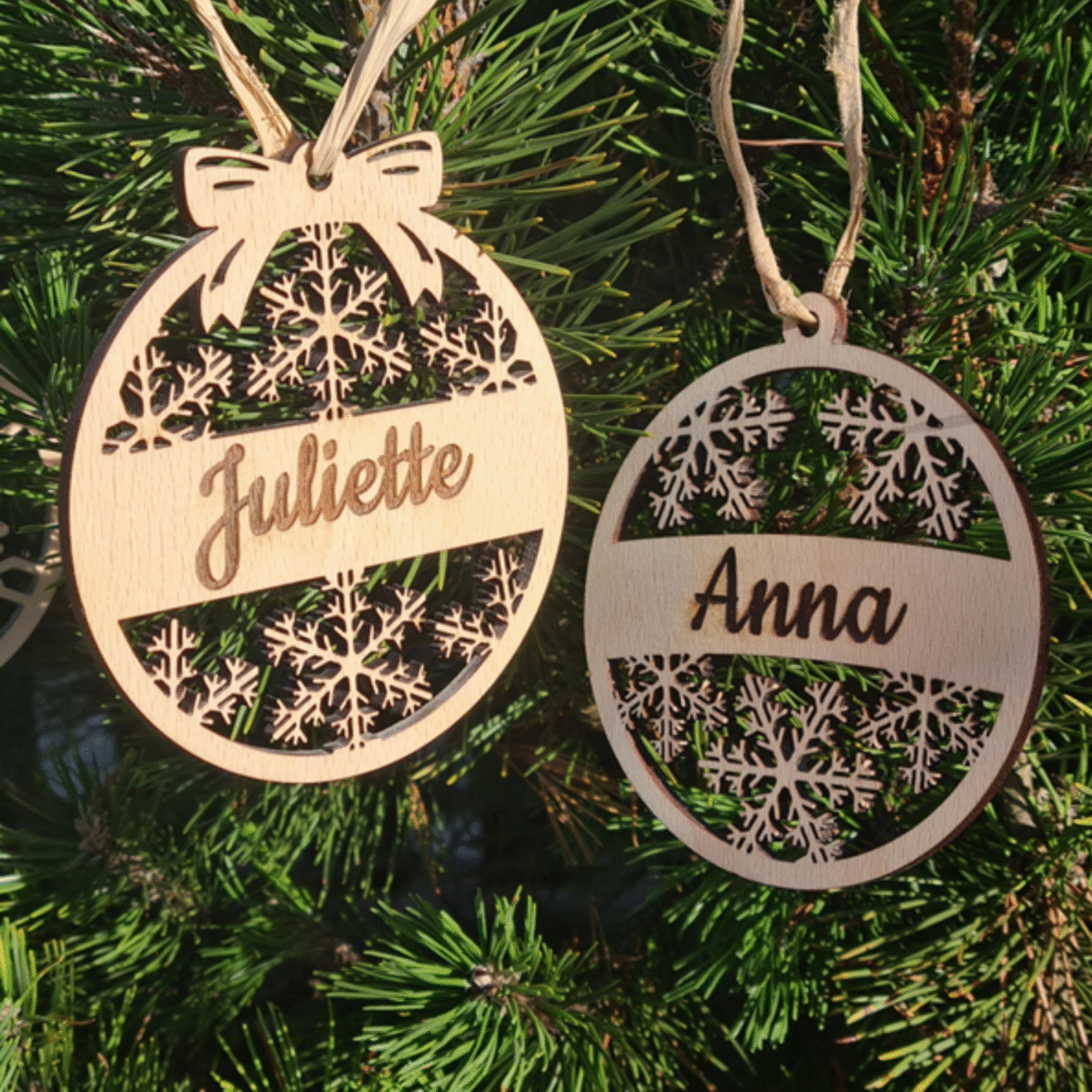 Collection de six boules de Noël personnalisées en bois suspendues aux branches d'un sapin, montrant divers motifs de flocons et décorations avec des prénoms incluant "Juliette" et "Anna"