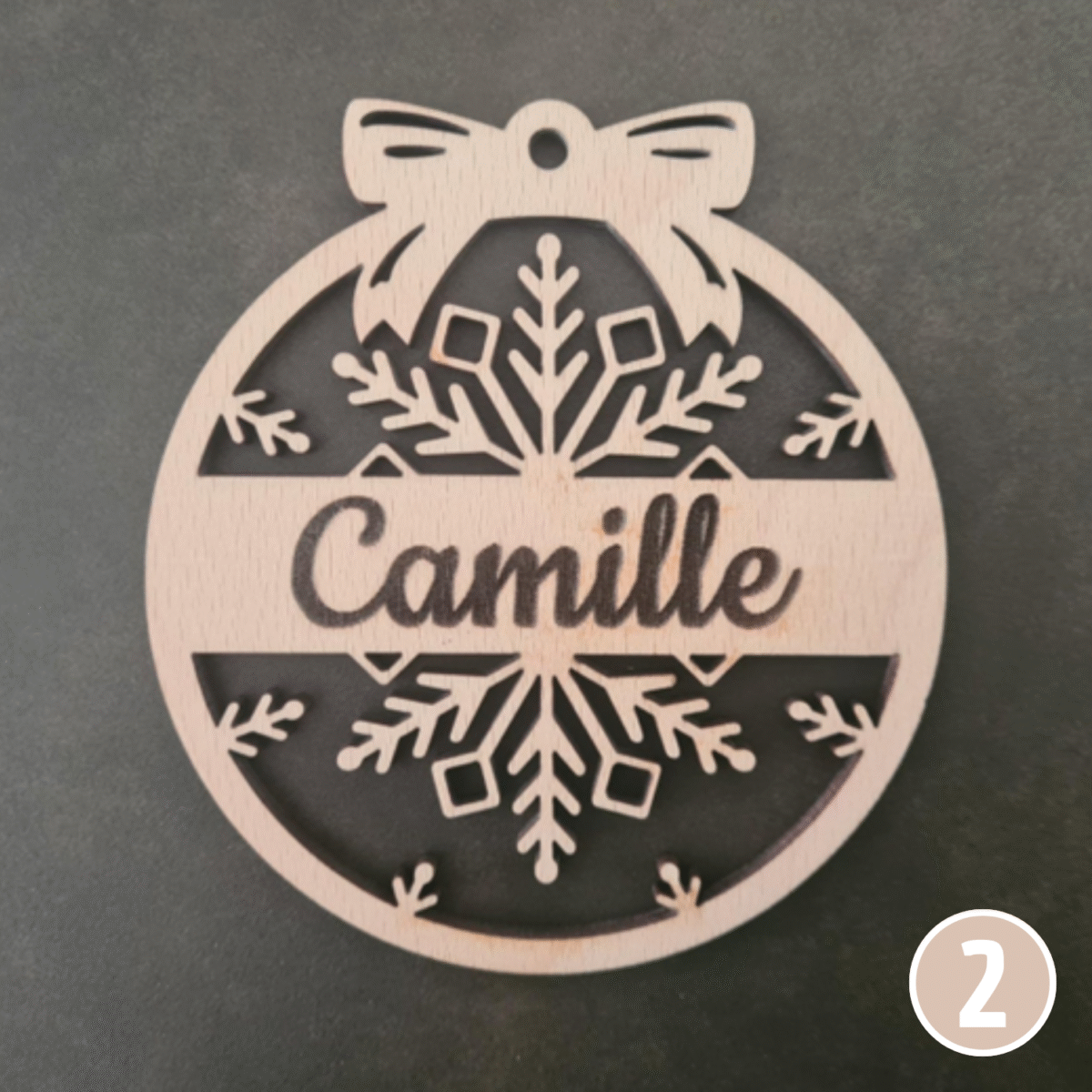 Boule de Noël personnalisée en bois avec le prénom "Camille", ornée d'un nœud décoratif en haut et de motifs flocons de neige ajourés tout autour du design circulaire