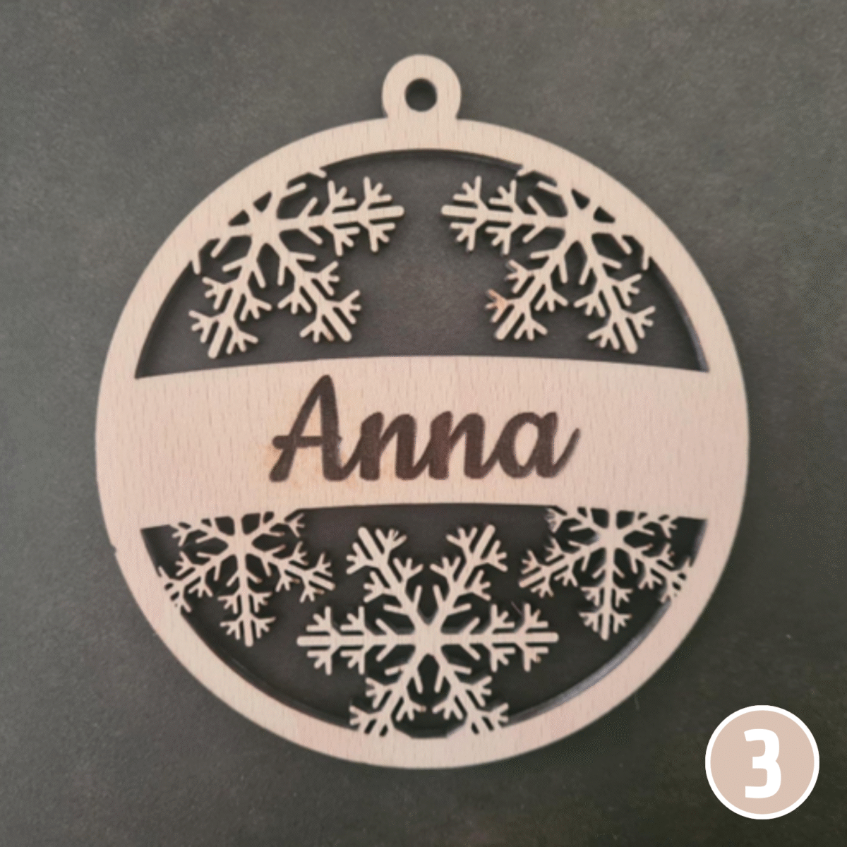 Décoration de Noël ronde en bois personnalisée avec le prénom "Anna", présentant de délicats flocons de neige découpés au laser dans les sections supérieure et inférieure avec anneau de suspension