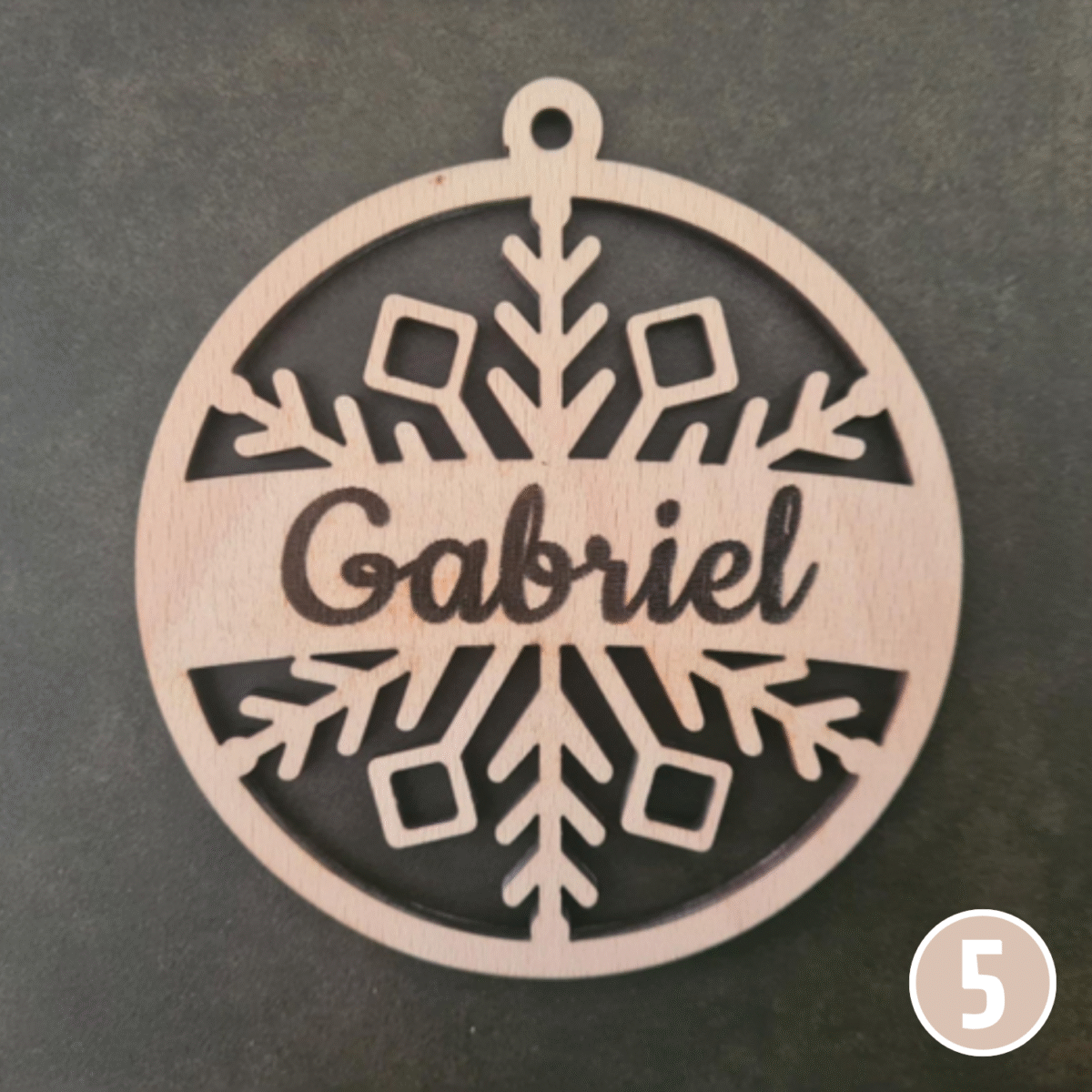 Boule de Noël personnalisée en bois affichant le prénom "Gabriel" avec d'élégants motifs de flocons au-dessus et en-dessous du texte, forme circulaire avec anneau de suspension